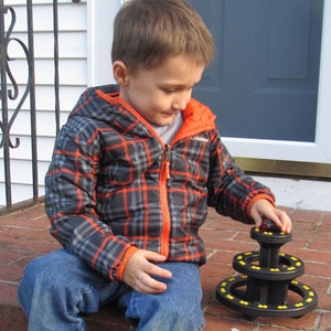 May include: A young boy wearing a plaid jacket and blue jeans plays with a black and yellow stacking toy. The toy has three tiers and is made of plastic.