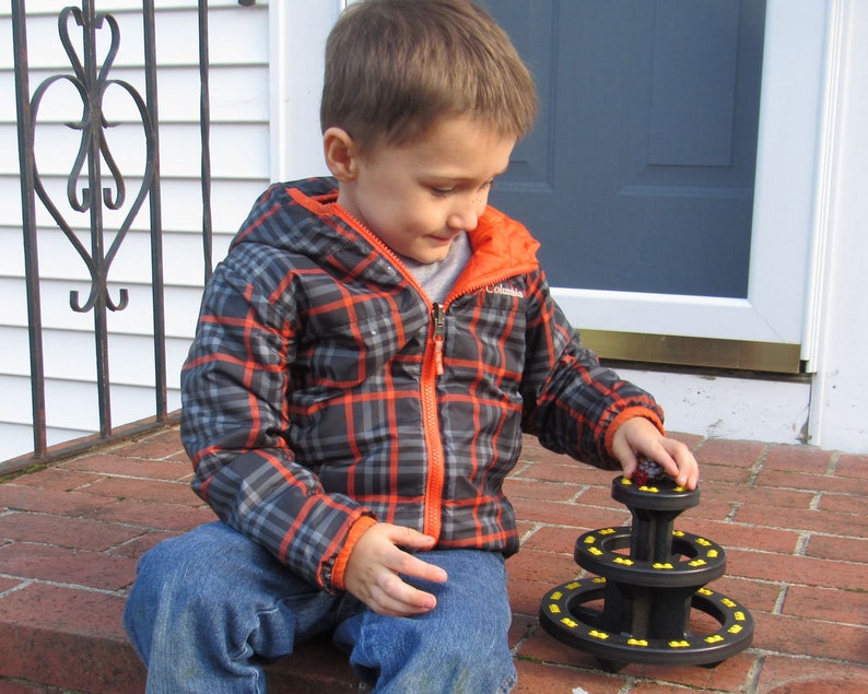 May include: A young boy wearing a plaid jacket and blue jeans plays with a black and yellow stacking toy. The toy has three tiers and is made of plastic.