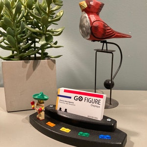Puede incluir: Un porta tarjetas de visita de pl&aacute;stico negro con una figura de Lego de un hombre con un sombrero y maracas. El soporte tiene un arco&iacute;ris de ladrillos Lego en la parte inferior y una tarjeta de visita con el texto "Go Figure Displays" y la informaci&oacute;n de contacto.