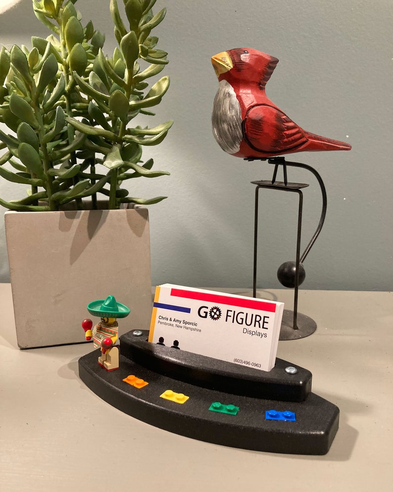 Puede incluir: Un porta tarjetas de visita de pl&aacute;stico negro con una figura de Lego de un hombre con un sombrero y maracas. El soporte tiene un arco&iacute;ris de ladrillos Lego en la parte inferior y una tarjeta de visita con el texto "Go Figure Displays" y la informaci&oacute;n de contacto.