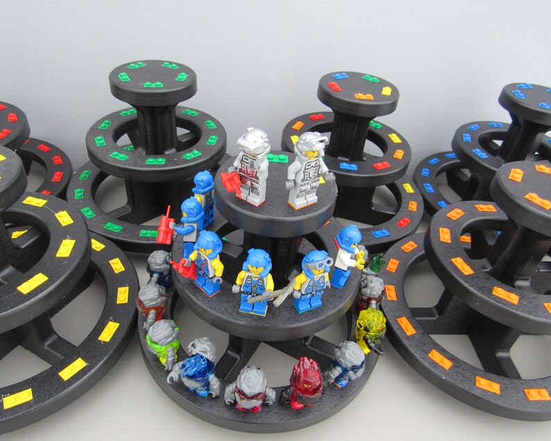 May include: A collection of small, colorful, plastic figurines arranged on a tiered display stand. The figurines are all wearing different colored helmets and have various accessories. The stand is made of black plastic and has a circular base.
