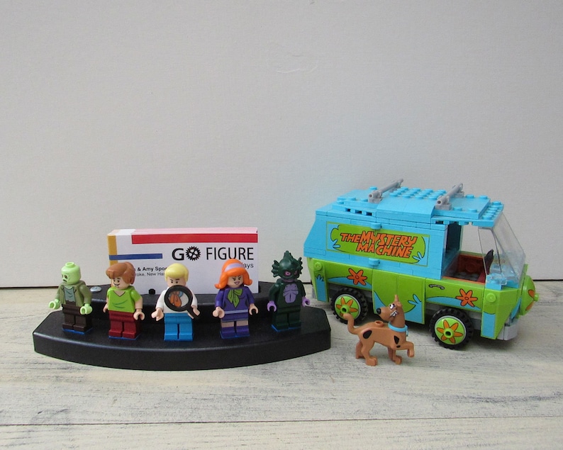 Puede incluir: Un conjunto de seis minifiguras de Scooby-Doo, incluyendo Scooby-Doo, Shaggy, Velma, Daphne, Fred y un monstruo, expuestas en un soporte negro. La furgoneta Mystery Machine tambi&eacute;n est&aacute; incluida, con las palabras "The Mystery Machine" impresas en el lateral.
