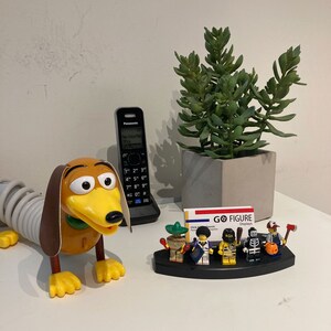 Puede incluir: Un perro salchicha de juguete con cola de resorte, un tel&eacute;fono negro, una planta suculenta en una maceta gris y un porta tarjetas de visita con una tarjeta de visita que dice "GO FIGURE Displays" y cinco figuras en miniatura.
