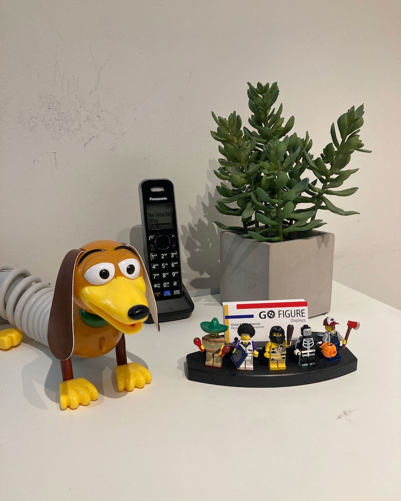 Puede incluir: Un perro salchicha de juguete con cola de resorte, un tel&eacute;fono negro, una planta suculenta en una maceta gris y un porta tarjetas de visita con una tarjeta de visita que dice "GO FIGURE Displays" y cinco figuras en miniatura.