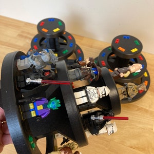May include: A multi-level rotating display stand with a black base and colorful accents. The stand features various Star Wars action figures, including a Stormtrooper, a Darth Vader, and a Rebel pilot. The figures are arranged in a circular pattern on the stand, creating a dynamic and eye-catching display.