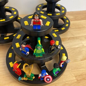 May include: A black tiered display stand with yellow accents holds a collection of superhero and cartoon character figurines. The figurines include Wonder Woman, Superman, a green monster, a red and blue superhero, a red and blue cartoon character, a green cartoon character, a red cartoon character, and a blue cartoon character.