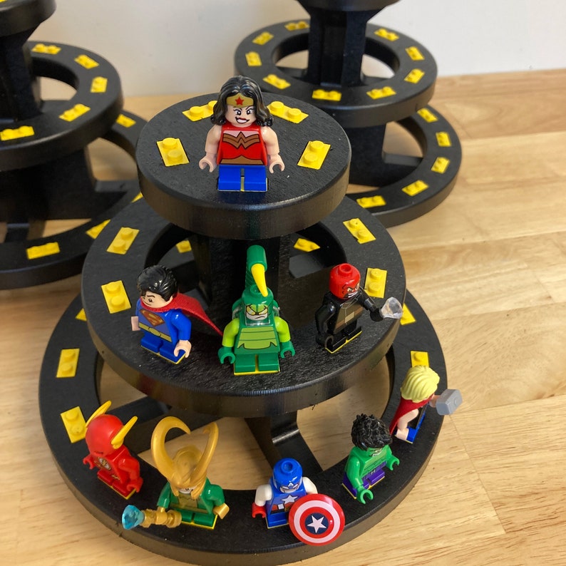 May include: A black tiered display stand with yellow accents holds a collection of superhero and cartoon character figurines. The figurines include Wonder Woman, Superman, a green monster, a red and blue superhero, a red and blue cartoon character, a green cartoon character, a red cartoon character, and a blue cartoon character.