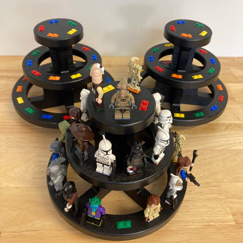 May include: A three-tiered black display stand with colorful rings, holding a collection of Star Wars minifigures. The figures include characters like Darth Vader, Stormtroopers, and a variety of alien characters.