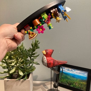 Puede incluir: Un expositor de madera negro curvado sostiene una colecci&oacute;n de minifiguras Lego de colores. Las minifiguras est&aacute;n dispuestas en fila, mirando hacia abajo, e incluyen un jardinero, un chef, un vaquero, un caballero y un granjero. El expositor est&aacute; siendo sostenido por una mano, y una figurita de p&aacute;jaro de madera roja es visible en el fondo.