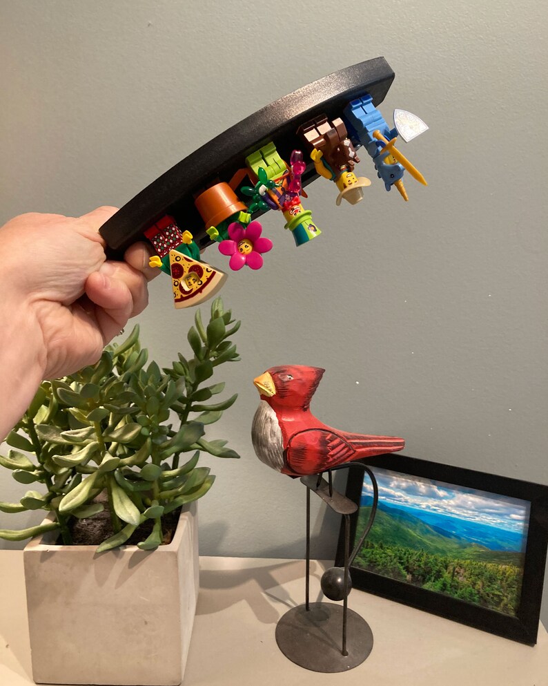 Puede incluir: Un expositor de madera negro curvado sostiene una colecci&oacute;n de minifiguras Lego de colores. Las minifiguras est&aacute;n dispuestas en fila, mirando hacia abajo, e incluyen un jardinero, un chef, un vaquero, un caballero y un granjero. El expositor est&aacute; siendo sostenido por una mano, y una figurita de p&aacute;jaro de madera roja es visible en el fondo.