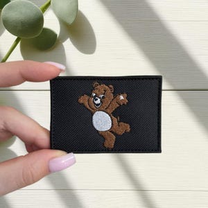 Op de afbeelding: Geborduurde patch met een bruine beer met een witte buik en een lachend gezicht, tegen een zwarte rechthoekige achtergrond. De beer staat in een speelse pose met uitgestrekte armen. De patch wordt door een persoon vastgehouden.