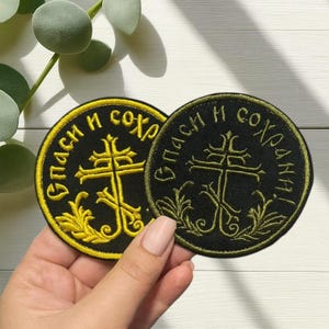 Peut inclure: Deux écussons ronds brodés avec le texte "Спаси и сохрани!" en or et vert olive. Chaque écusson présente un motif de croix avec des accents floraux. Les écussons sont sur un fond noir.