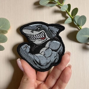 Puede incluir: Parche bordado con un tiburón musculoso flexionando su brazo. El tiburón es gris con boca blanca y lengua roja, y está delineado en negro. El parche es probablemente para ropa o accesorios.