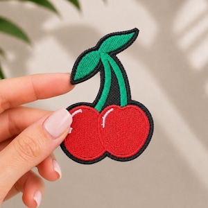 Puede incluir: Un parche bordado con forma de cerezas que presenta dos frutos circulares de color rojo brillante con reflejos blancos y un tallo verde con dos hojas, todo delineado con un borde negro grueso para aplicar sobre telas.