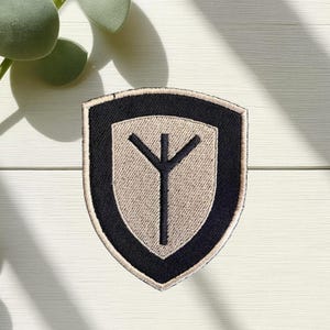 Peut inclure: Un écusson en forme de bouclier avec une bordure noire et un intérieur beige. L'écusson présente un symbole noir ressemblant à un arbre ou un trident stylisé. L'écusson est probablement un emblème ou un insigne.