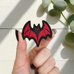 Op de afbeelding: Geborduurde vleermuispatch met een zwarte omtrek en een rode binnenkant. De vleermuis heeft een gedetailleerd vleugelontwerp. De patch wordt door een hand omhoog gehouden.