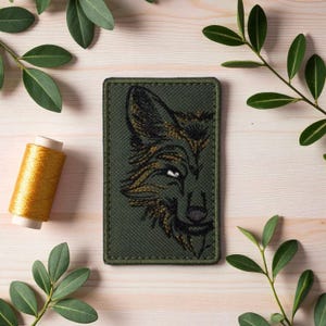 May include: A rectangular olive green embroidered patch featuring a detailed wolf head design in black and gold. The patch is surrounded by a dark green border. A spool of gold thread and green leaves are in the background.