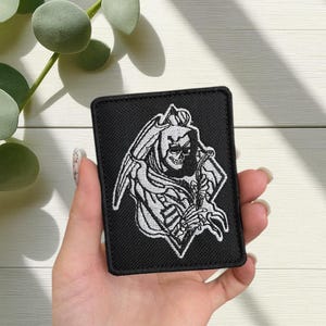 Può includere: Toppa rettangolare nera con un disegno del Tristo Mietitore ricamato in bianco. Il Mietitore è raffigurato con un volto scheletrico, un mantello con cappuccio e una falce. La toppa è per abbigliamento o accessori.