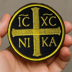 May include: Round black patch with a gold cross design and the Greek letters IC XC NI KA. The patch has a gold border and is likely an embroidered emblem.