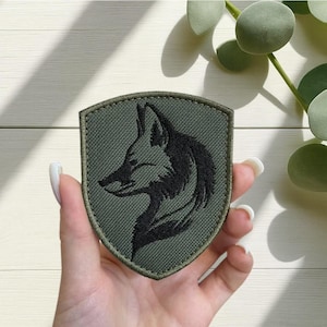 Könnte beinhalten: Ein olivgrüner, gestickter Aufnäher in Schildform. Der Aufnäher zeigt eine schwarze Silhouette eines Wolfskopfes im Profil. Der Aufnäher wird in einer Hand vor einem hellen Hintergrund mit grünem Laub gehalten.