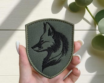 May include: An olive green embroidered patch in a shield shape. The patch features a black silhouette of a wolf's head in profile. The patch is held in a hand against a light background with green foliage.