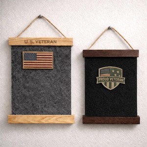 May include: Two felt wall hangings with wooden frames and rope hangers. One is grey with a U.S. flag patch and the text "U.S. VETERAN." The other is black with a "PROUD VETERAN" patch.