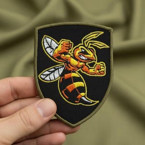 May include: Embroidered patch featuring a stylized, muscular wasp in orange, yellow, and black, with white wing details. The wasp is depicted in a dynamic, aggressive pose. The patch has a black background and a green border.