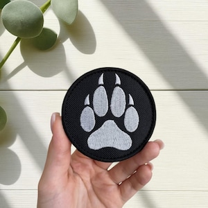 May include: A round, black patch featuring a white paw print design. The patch is embroidered with a detailed paw print, showcasing the claws and pads. The patch is held in a hand against a white wooden background.