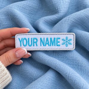 May include: A rectangular embroidered patch with a white background and a light gray border. The text "YOUR NAME" is stitched in bright blue capital letters, followed by a blue snowflake icon on the right side of the patch.