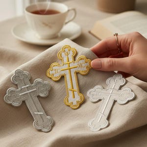 Può includere: Toppe ricamate a forma di croce in argento, oro e bianco, con dettagli intricati. Le croci sono esposte su un tessuto beige, con una tazza di tè e un libro sullo sfondo. Le toppe sono probabilmente per scopi religiosi o decorativi.