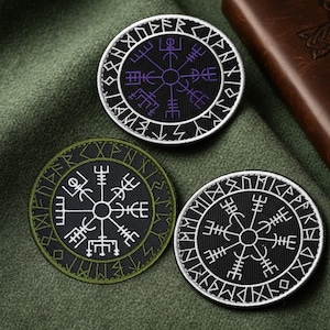 May include: Three circular embroidered patches with Viking symbols. The patches feature a central symbol surrounded by runes, in black, white, purple, and green. The patches are on a green fabric background.