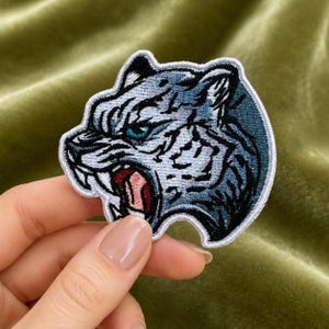 Puede incluir: Un parche bordado que muestra la cabeza de un tigre gruñendo en tonos azules, blancos y negros. El tigre tiene un ojo azul brillante, la boca abierta de color rosa con dientes visibles y un borde blanco grueso que perfila todo el diseño.