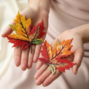 Pode incluir: Duas aplicações bordadas em forma de folha de bordo em cores outonais: vermelho, laranja, amarelo e verde. As folhas são seguradas em mãos abertas, mostrando as costuras detalhadas e os tons vibrantes. As aplicações são provavelmente para costura ou artesanato.