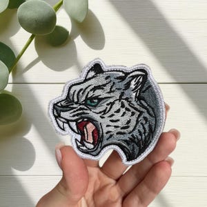 Pode incluir: Patch bordado com a cabeça de um tigre rosnando. O patch tem uma borda branca, detalhes cinza e preto e uma boca vermelha. Os olhos do tigre são verdes. O patch é segurado em uma mão.