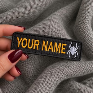 May include: A rectangular black embroidered patch featuring the yellow text YOUR NAME and a white spider graphic on the right side. The patch has a black stitched border and is designed for attachment to fabric items as a personalised identifier.