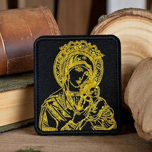 Puede incluir: Un parche cuadrado negro con una imagen bordada en oro de la Virgen María sosteniendo al niño Jesús. El diseño incluye un halo y una corona, con costuras detalladas. El parche es probablemente para ropa o accesorios.