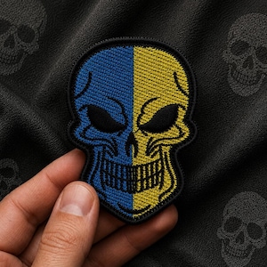 Op de afbeelding: Een geborduurde schedelpatch met een gespleten ontwerp met een levendige blauwe linkerkant en een heldergele rechterkant. De schedel heeft zwart geborduurde oogkassen, een zwarte neusholte en een zwarte grijns, omlijnd met zwart garen.