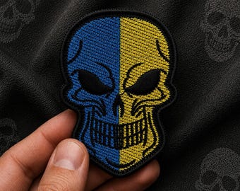 Op de afbeelding: Een geborduurde schedelpatch met een gespleten ontwerp met een levendige blauwe linkerkant en een heldergele rechterkant. De schedel heeft zwart geborduurde oogkassen, een zwarte neusholte en een zwarte grijns, omlijnd met zwart garen.
