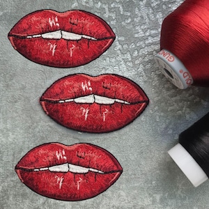 Puede incluir: Tres parches bordados de labios rojos con dientes blancos sobre una superficie gris texturizada. Un carrete de hilo rojo y un carrete de hilo negro son visibles en el fondo, lo que sugiere un tema de costura o artesanía.