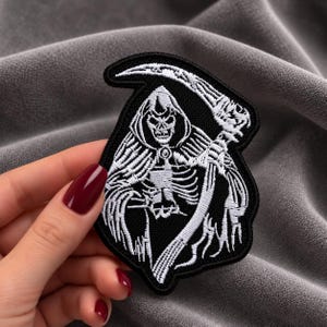 Peut inclure: Un écusson brodé en noir et blanc représentant une faucheuse vêtue d'une robe à capuche et tenant une grande faux. Le motif est réalisé en fil blanc sur un fond noir uni avec une bordure noire épaisse entourant la forme de l'écusson.
