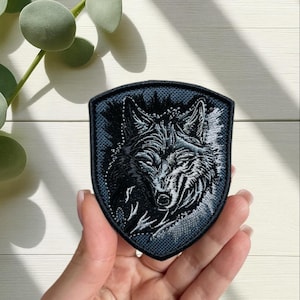 May include: A shield-shaped patch featuring a detailed embroidered wolf head in shades of black and gray. The wolf's snarling expression and fur texture are prominent. The patch has a dark blue border and is likely for clothing or accessories.