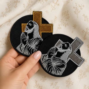 May include: Two circular embroidered patches featuring a figure with hands clasped in prayer against a black background. One patch includes a gold cross, while the other features a silver cross. Both designs show a detailed profile of a bearded figure.