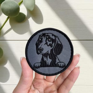 May include: Round embroidered patch depicting a black and tan dachshund dog. The patch has a grey background and a black border. The dog's head and paws are visible. The patch is held in a hand.