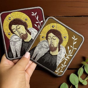 Puede incluir: Dos parches bordados con una figura religiosa con aureola dorada, cabello castaño y barba. Un parche tiene un fondo burdeos, el otro un fondo marrón. Ambos tienen alas bordadas en blanco y un diseño floral blanco.