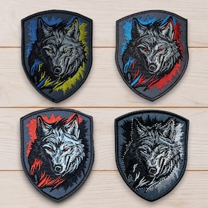 Könnte beinhalten: Vier gestickte Aufnäher mit Wolfsmotiv. Jeder Aufnäher zeigt einen Wolfskopf in Schildform. Das Fell des Wolfes ist grau, mit roten, blauen und gelben Akzenten. Die Aufnäher liegen auf einer hellen Holzmaserung.
