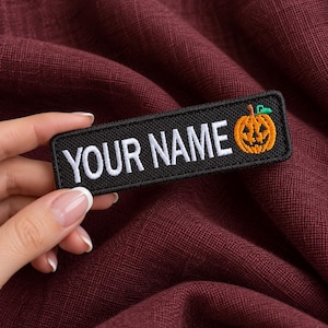 Op de afbeelding: Een rechthoekige zwarte geborduurde patch met de witte tekst YOUR NAME in hoofdletters, gevolgd door een oranje jack-o'-lantern pompoenpictogram met een groene steel aan de rechterkant, afgewerkt met een zwarte gestikte rand.