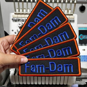 K&ouml;nnte beinhalten: Eine Reihe schwarzer rechteckiger Aufn&auml;her mit dem Text "Fam-Dam" in Blau gestickt. Jeder Aufn&auml;her hat einen orangefarbenen Rand. Die Aufn&auml;her werden vor einer Stickmaschine gehalten.