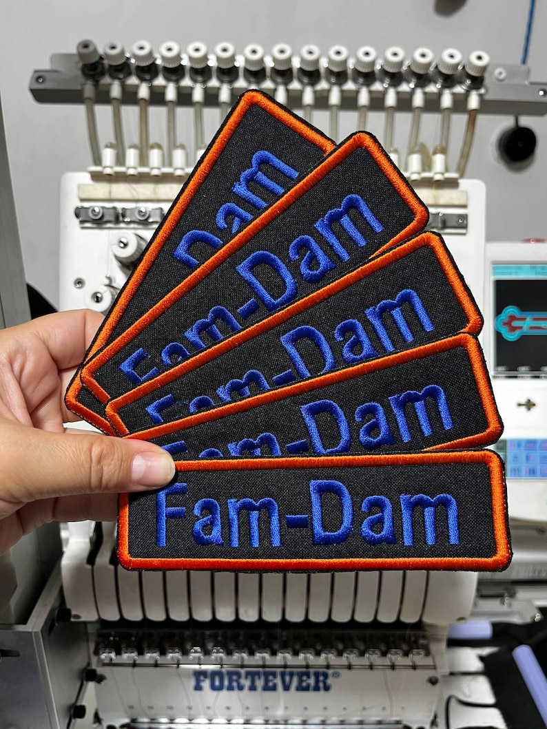 K&ouml;nnte beinhalten: Eine Reihe schwarzer rechteckiger Aufn&auml;her mit dem Text "Fam-Dam" in Blau gestickt. Jeder Aufn&auml;her hat einen orangefarbenen Rand. Die Aufn&auml;her werden vor einer Stickmaschine gehalten.