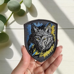 Peut inclure: Écusson brodé représentant une tête de loup détaillée avec des yeux jaunes, sur un fond en forme de bouclier. Le motif intègre des accents bleus et jaunes, créant un contraste saisissant sur un fond gris. L'écusson est tenu dans une main.