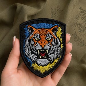 Op de afbeelding: Een schildvormige geborduurde patch met een gedetailleerde tijgerkop met oranje, witte en zwarte vacht. De tijger heeft een grommende uitdrukking met een rode bek. De achtergrond bevat blauwe en gele stiksels op een zwarte rand.